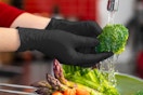 Two hands wearing black nitrile gloves wash broccoli under a faucet. Other veggies are nearby.