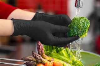 Two hands wearing black nitrile gloves wash broccoli under a faucet. Other veggies are nearby.