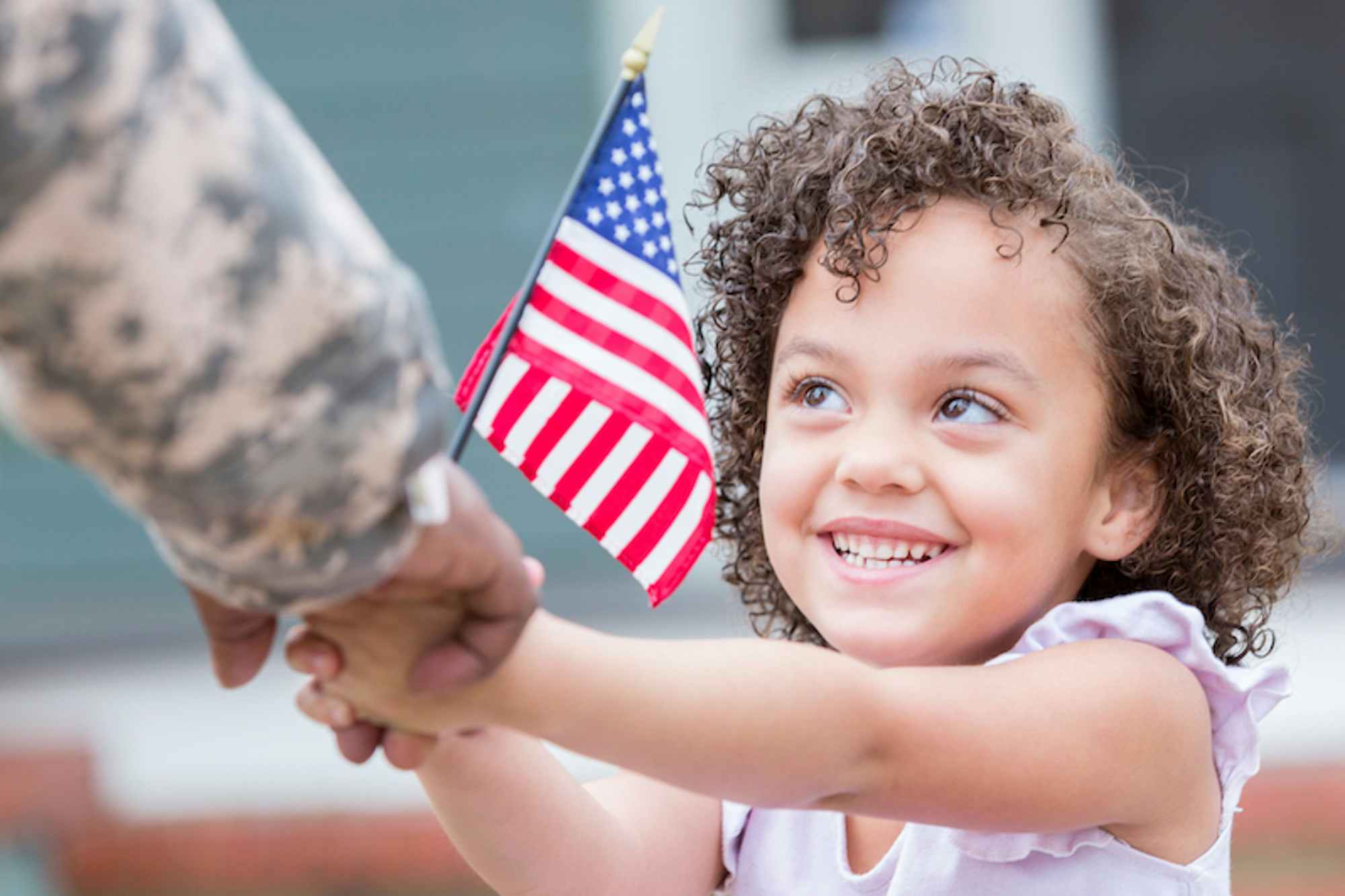 A little girl smiles up at her military dad while holding his hand. She is also holding a small US flag.
