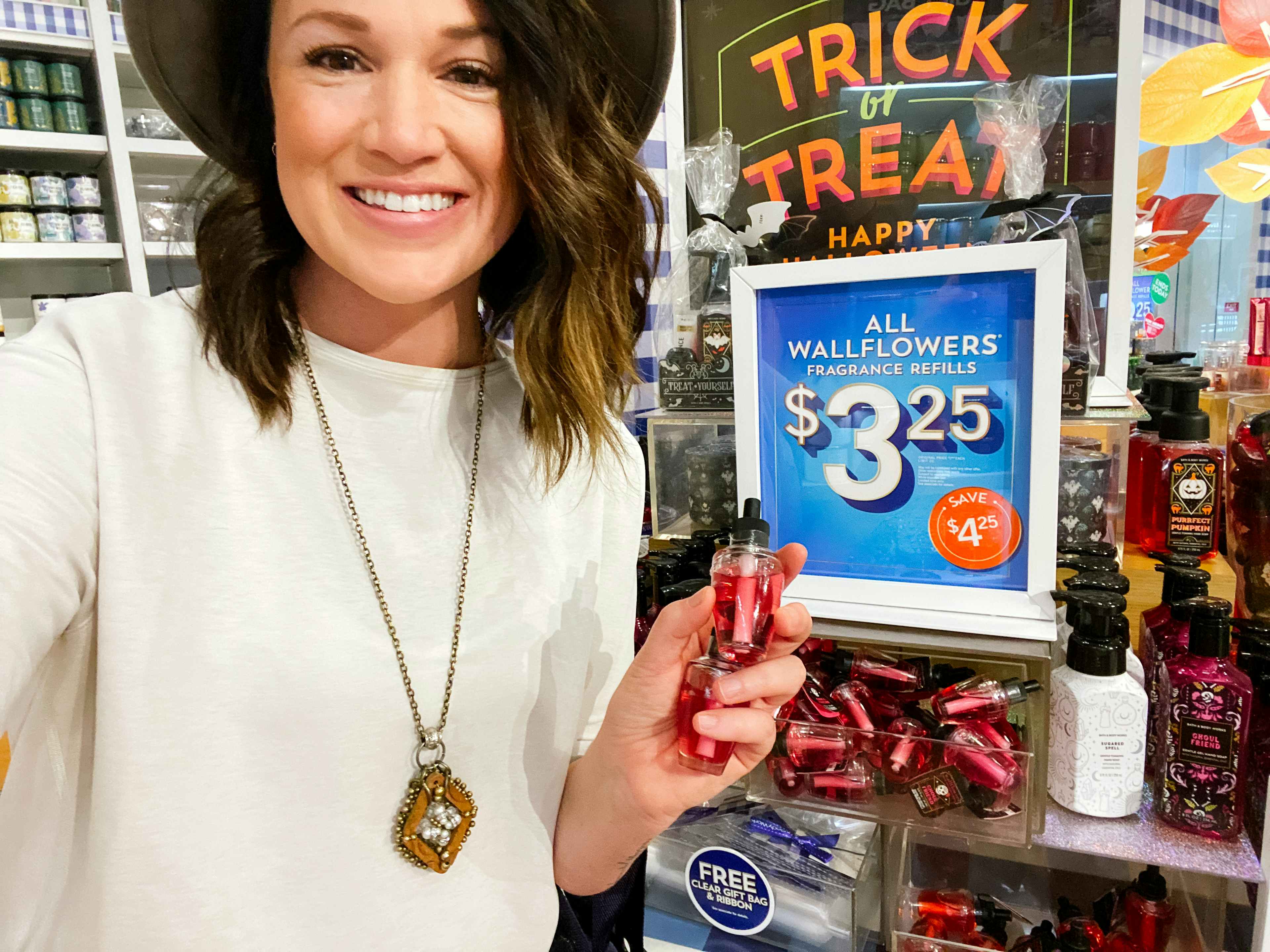 A woman smiling and holding up a Bath & Body Works wallflower refill in front of a sign that reads, "All wallflowers fragrance refills $3...