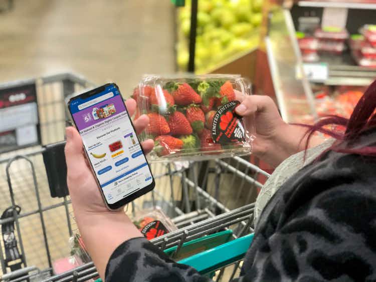 A person has the Kroger app on her phone while shopping and holding strawberries.