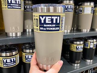 A person's hand holding up a beige Yeti Rambler 20oz tumbler in front of a shelf of more of the same tumbler at Dick's Sporting Goods.