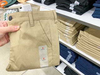 A person's hand holding a pair of children's school uniform pants next to a shelf of more pants at Old Navy.