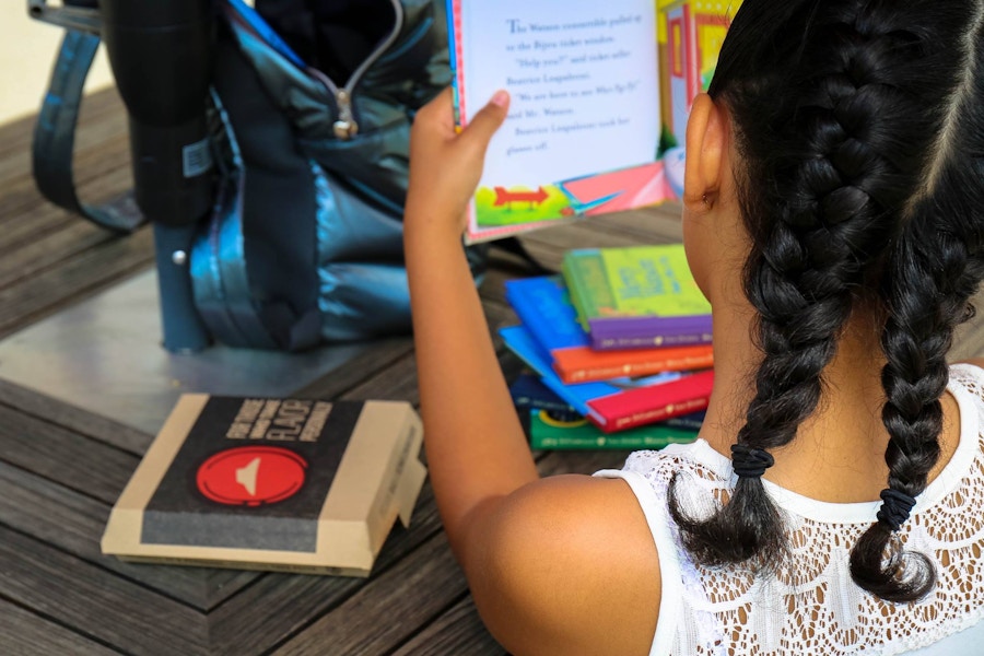 A child reading a book with a backpack, other books, and a pizza hut kids pizza box next to her.