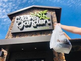 Person holding an Olive Garden to-go bag in front of an Olive Garden restaurant.