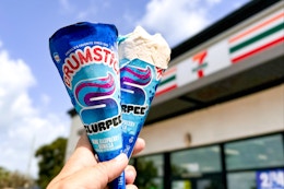 Two slurpee drumsticks being held up in front of a 7 eleven store