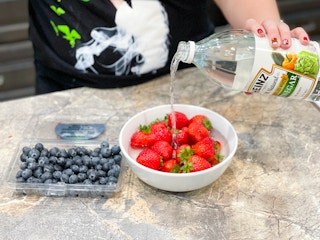 A person pouring Heinz vinegar into a bowl of strawberries sitting on a counter next to a container of blueberries.