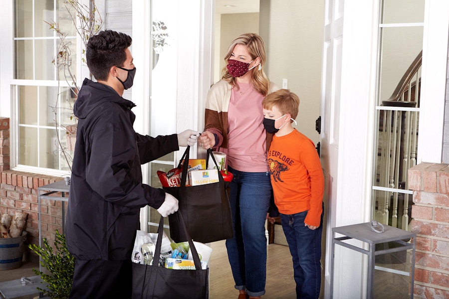 A man wearing a mask and gloves handing groceries to a woman and young boy, also wearing masks, at the front door of a house.