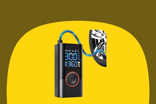 A black rectangular tire inflator with large blue LCD numbers and a large round power-mode button bordered in red, attached to a tire.