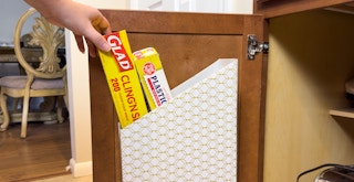 Someone taking some plastic wrap out of a magazine holder attached to the inside of a kitchen cabinet