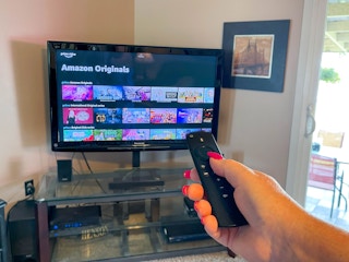A person's hand holding an Amazon Fire remote, aiming it at a TV displaying Prime Video's Amazon Originals.