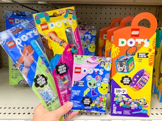A person's hand holding two LEGO Dots bracelet kits and accessories in front of a shelf with more LEGO Dots sets on the shelf in the ...