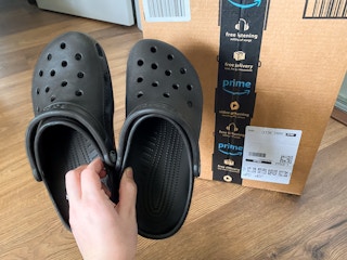 Someone holding up a pair of Crocs next to an Amazon box
