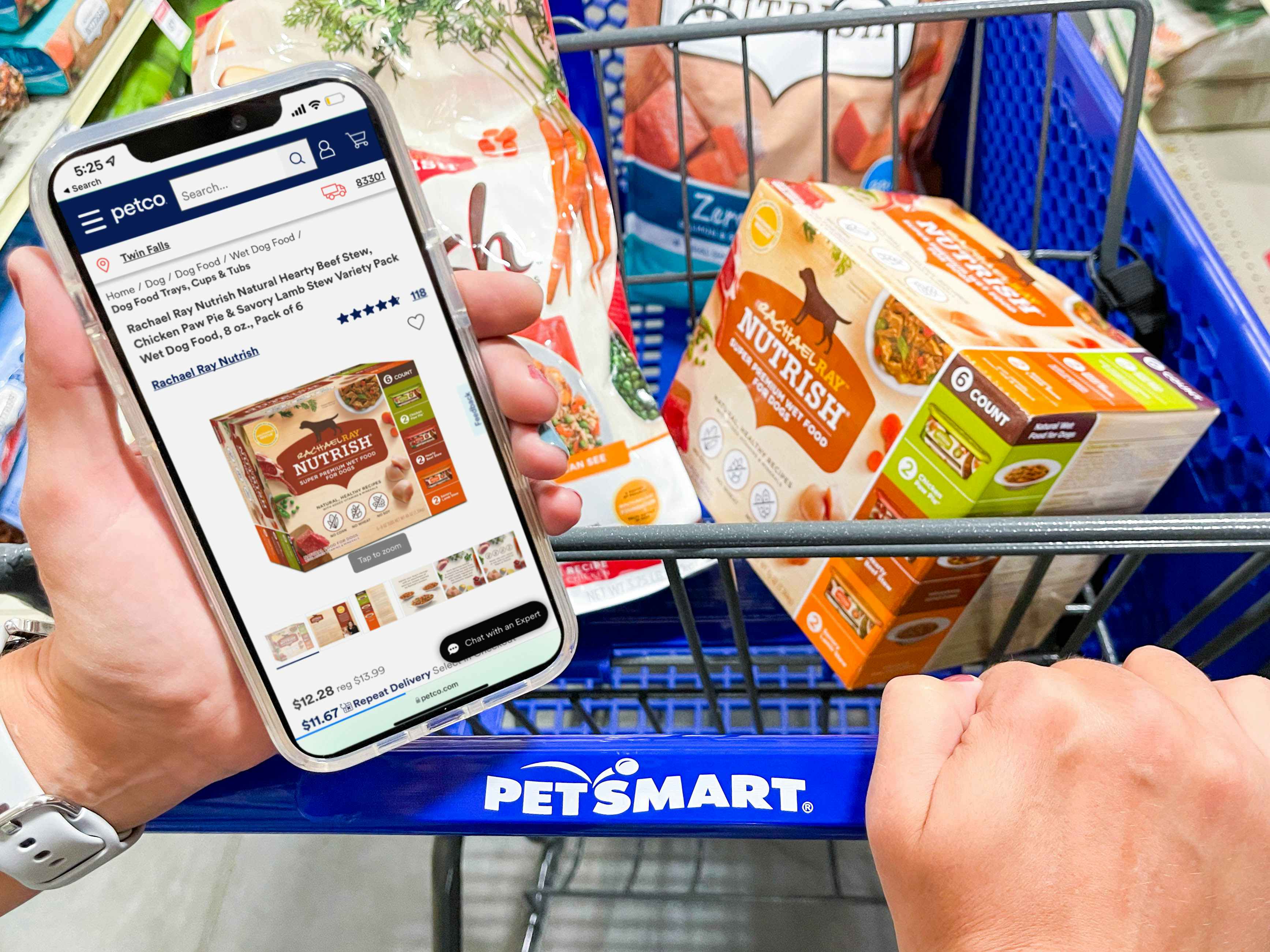 A person's hands, one holding a cellphone with the Petco website open to a product, and the other hand pushing a PetSmart shopping cart w...