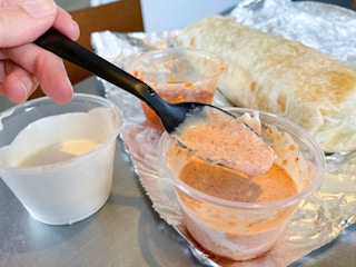 A black plastic spoon in a container filled with a pinkish colored sauce. Beside it are two sauce containers with sour cream and a hot sa...