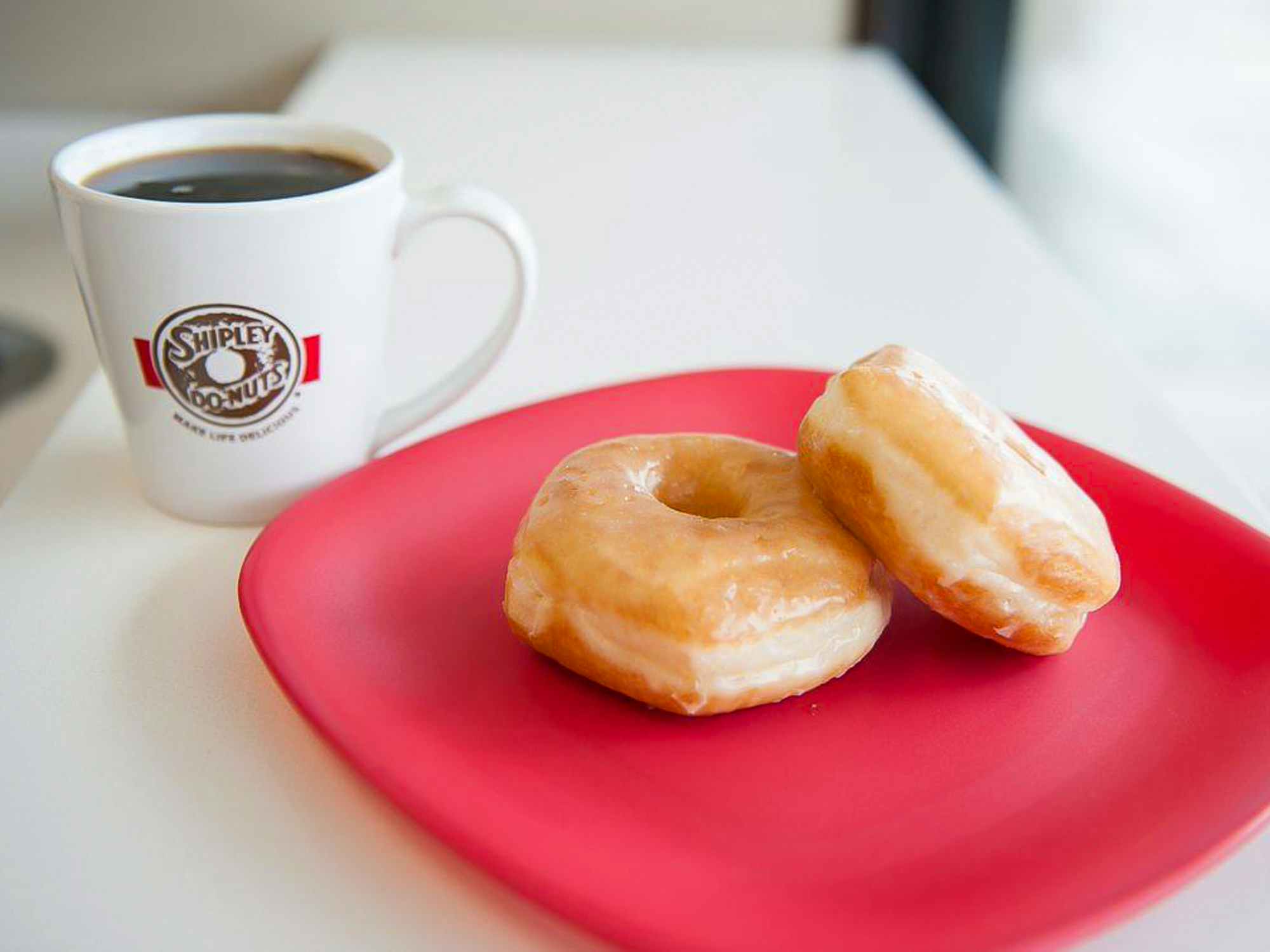 Some Shipley Do-nut doughnuts on a plate next to a cup of coffee