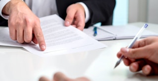 A person pointing to a signature line on a document while giving it over to someone holding a pen.