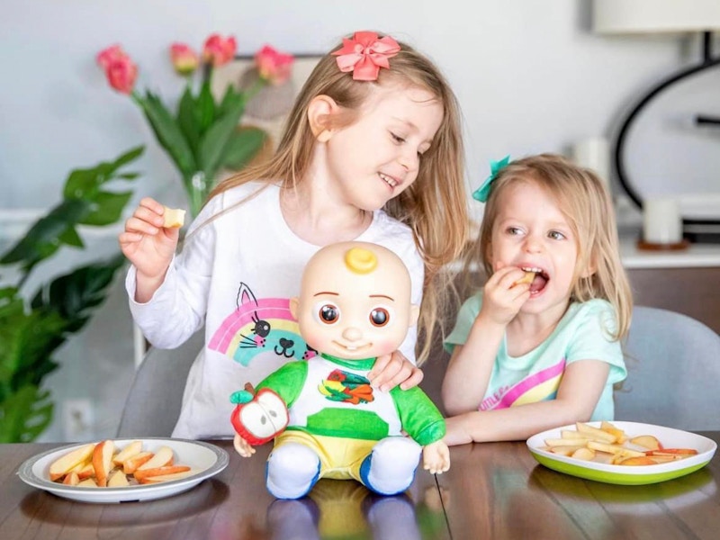 Two kids at a table in a dinning room, eating sliced apples with a Cocomelon doll between them.