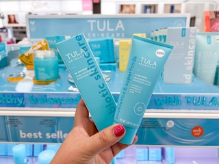 A hand holding a a boxed and unboxed product of Tula So Poreless Exfoliating Scrub in front of other Tula products