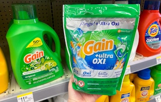 hand holding a bag of gain flings next to large size gain laundry detergent