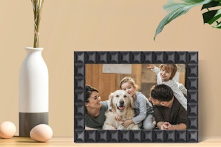 On a table in a beige room next to an artificial plant, a digital picture frame with a black pattern like inside out cubes has a happy famil