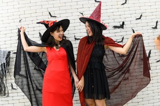 Two women dressed as witches smiling at each other in front of a white brick wall.