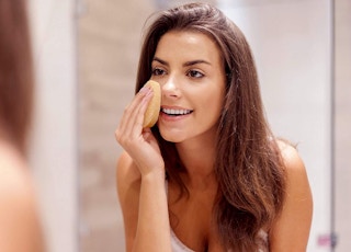 woman standing in front of mirror cleansing face with buf-puf sponge