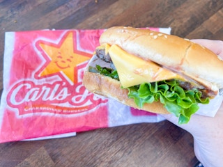 a hand holding a cheese burger from carls jr with a wrapper behind on table