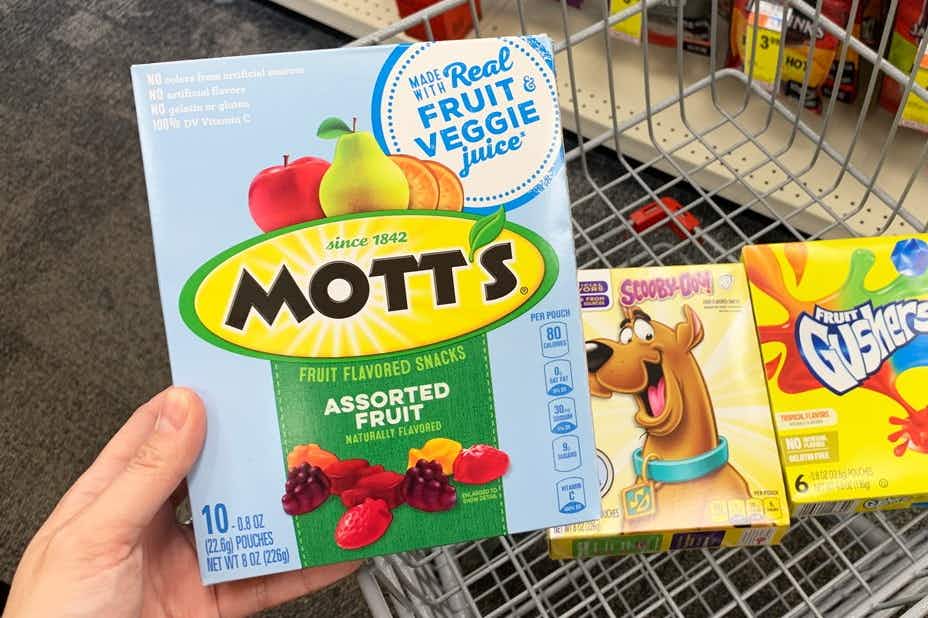 hand holding Motts fruit snacks with Betty crocker fruit snacks in a shopping cart in the background