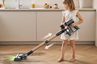 A girl child uses the Miuzzy cordless vacuum on a wood-look kitchen floor. The hands adusts for her height. The vac glows green for light.