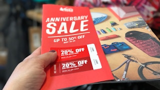 A person's hand holding up some REI Anniversary Sale coupons above a REI ad book.