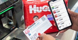 Ibotta app on a cell phone next to coupons and Huggies diapers at a Walmart checkout stand.