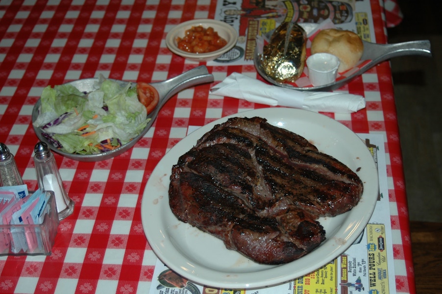 A steak on a plate, salad in a bowl, a baked potato in a bowl and a roll on a plate all sitting on a table.