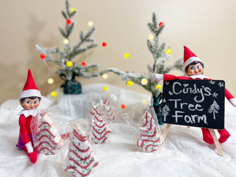 two elf on the shelf dolls with packaged cake trees and a cindy's tree farm chalk board sign 