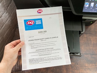 a person's hand holding a printed out egift card from dairy queen in front of a printer