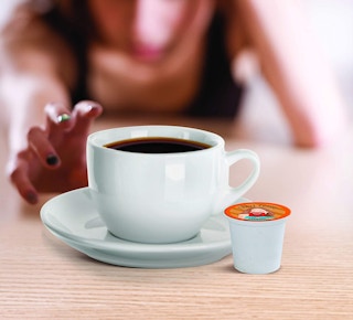 A hand reaching for a cup of coffee with a K-Cup next to it