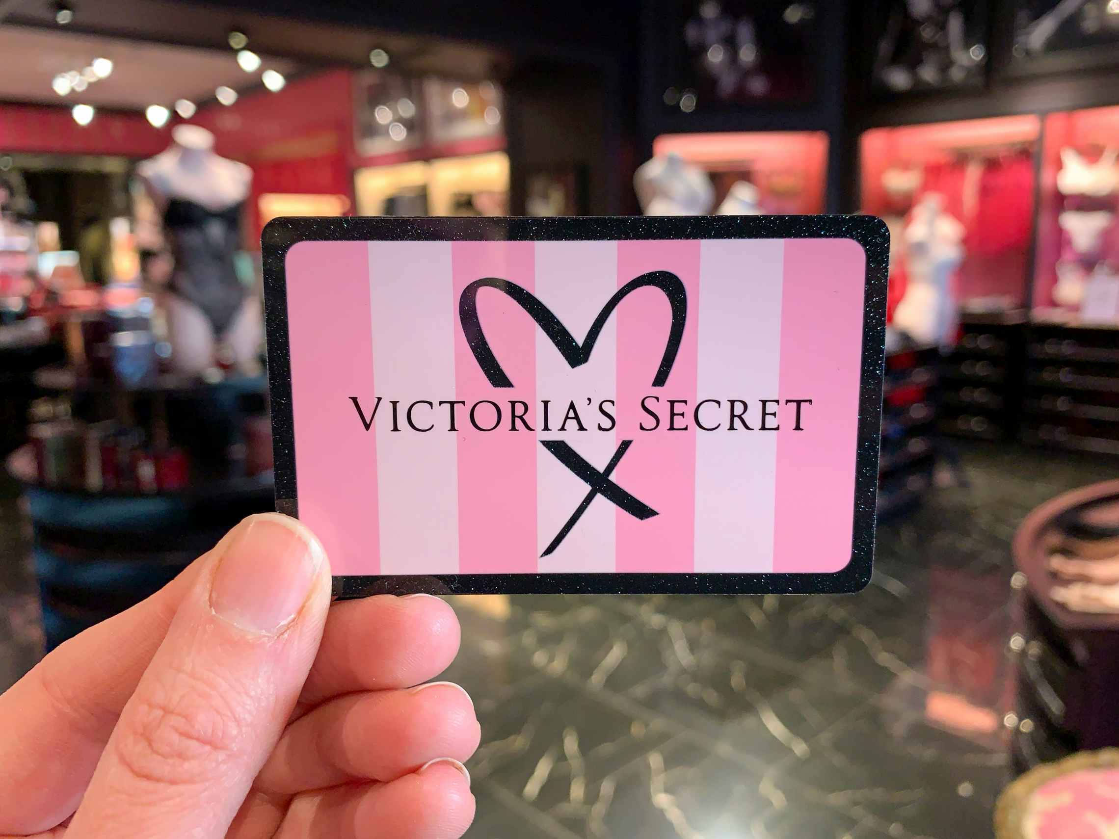 A person's hand holding a Victoria's Secret gift card inside a Victoria's Secret store.