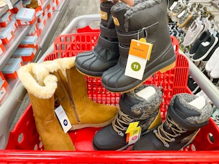 A person's hand holding a pair of children's boots above a Target shopping cart that has more children's boots in the basket, parked in ...