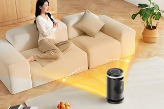 In a beige living room, a woman basks in the heat from her 1500-watt oscillating space heater.