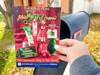 A person holding a Bath & Body Works coupon mailer next to a mail box