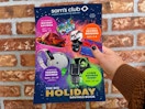 A person's hand holding up a Sam's Club Holiday Savings book in front of a brick wall.