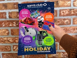 A person's hand holding up a Sam's Club Holiday Savings book in front of a brick wall.
