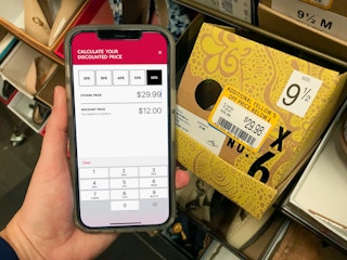 A person holding and iPhone next to a shoe box with a yellow clearance sticker on it. The cell phone screen displays the Discount ...