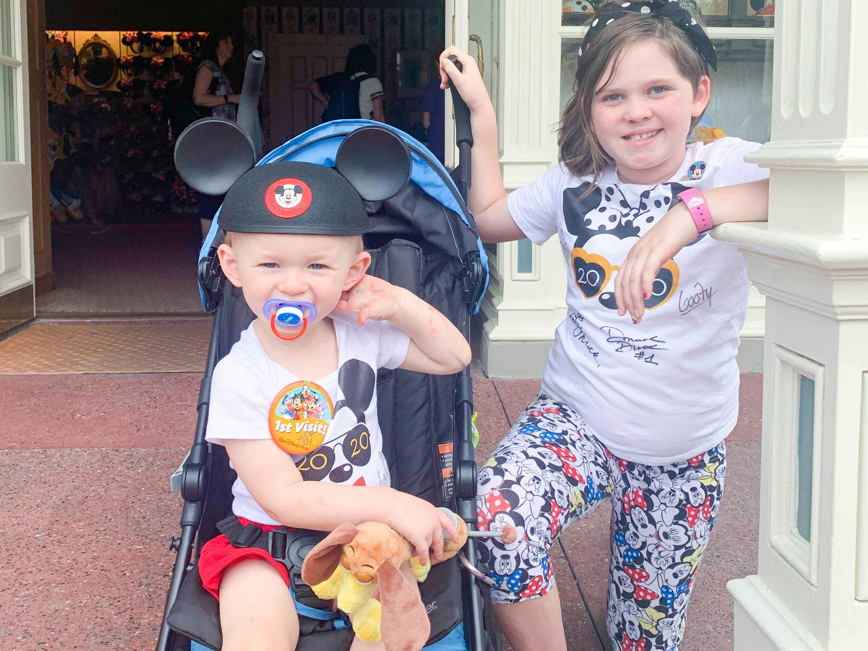 Two kids at Disney Worls, one sitting in a stroller wearing a 1st Visit pin and Mickey Mouse ears