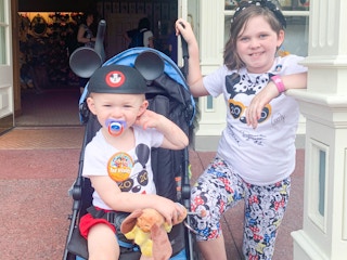 Two kids at Disney World, one sitting in a stroller wearing a 1st Visit pin and Mickey Mouse ears