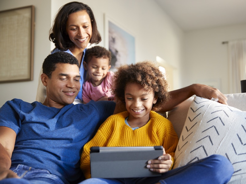 A family sitting together in the living room, all of them looking down at a tablet.