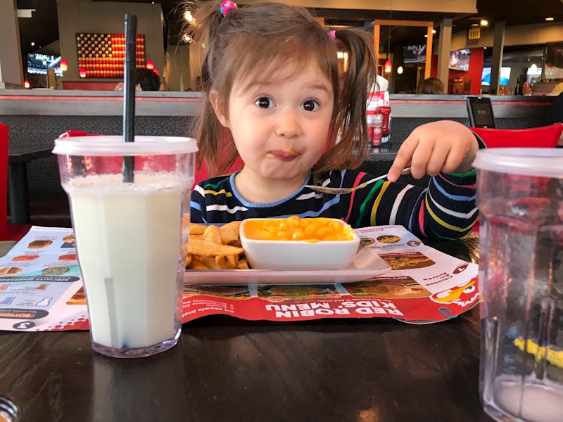 A child sitting in a Red Robin booth eating mac and cheese with a spoon, with a glass of milk in front of her.