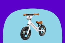 A white balance bike w. white frame, brown trim, and black 12-inch tires. It has a number 1 on a shield below the handlebars, and no pedals.