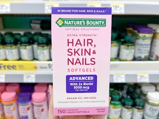 A box of Nature's Bounty Hair, Skin, & Nails held out by hand in front of a store shelf with other vitamin products sitting on it.