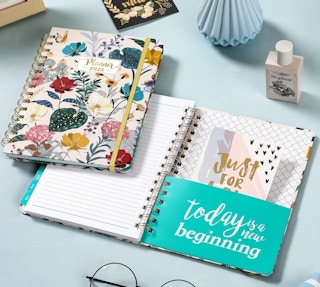 a closed planer with flowers on top of an open planner on a blue table